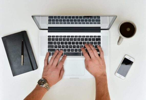 Hands of a man shown typing on a laptop computer whilst sitting at a table surrounded with a book cellphone and mug of coffee