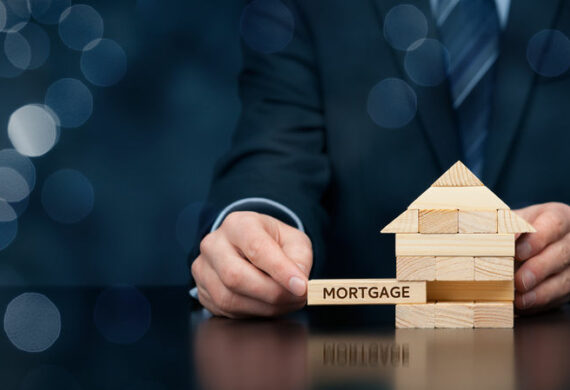 A small wooden house with the word mortgage displayed on a table by the hands of a man wearing a suit